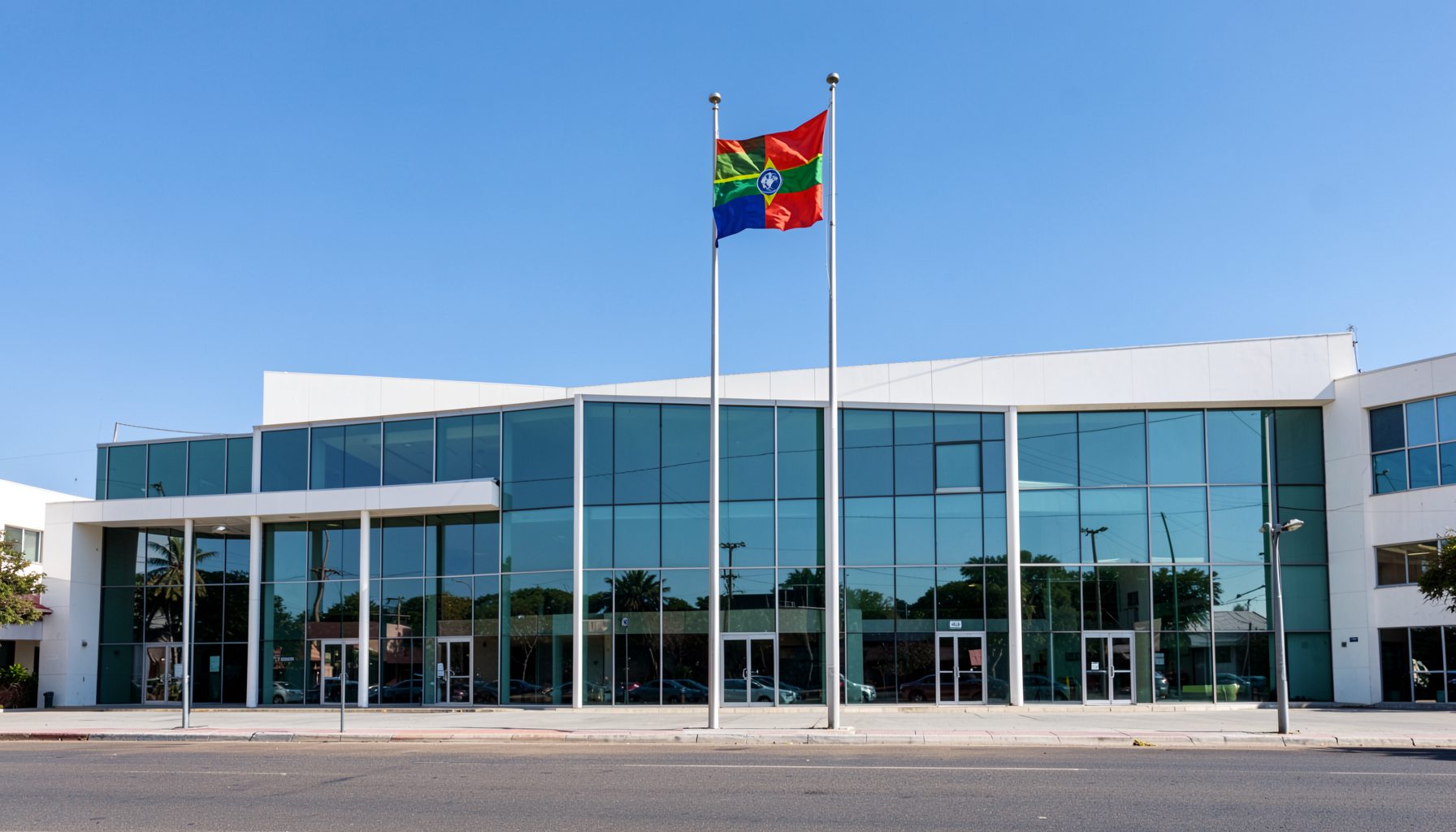 A wide shot of a modern civic building facade with clean lines, glass panels, and the Namibian flag flying prominently against a clear blue sky